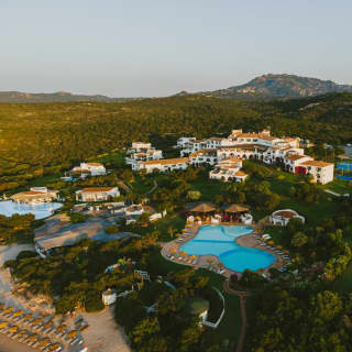 Aerial shot of Romazzino, white walls and blue pools illuminated by early sun, nestled in gardens between forest and beach.