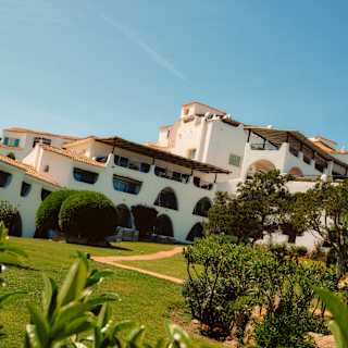 Angled image of Romazzino's white exterior with arch and rhombus windows and roof terraces, seen from lush green gardens.