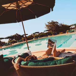A woman reads a magazine in the tub-backed poolside lounge chair, one arm flopped over the back, seen from behind.