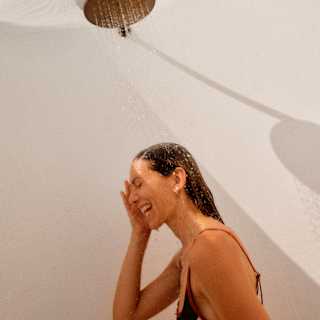 Low-angle view of a dark-haired woman in a bikini beneath a monsoon head al-fresco shower, seen from the chest up.