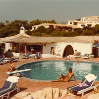 Historic honeyed Kodachrome photo of guests lounging with drinks by the sun-drenched resort pool, with a thatched bar area.