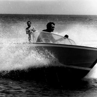 Historic black and white image of a man steering a speed boat, kicking up spray as he tows a water-skier in his wake.
