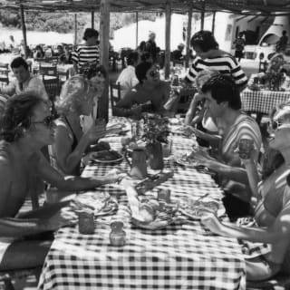Historic black and white image of the busy terrace, as guests lunch at sun-dappled tables, helped by waiters in striped tops.