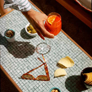 A hand places a Negroni cocktail between bowls of crisps, nuts, olives and a pair of sunglasses on a grey mosaic table top.