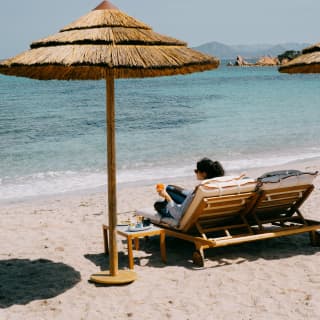 A dark-haired woman holds an orange juice as she reclines on one side of a twin sun-lounger next to a parasol on the beach.