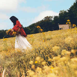 Low-angle image of a dark-haired woman in a white skirt and red top as she carries a basket of harvested flowers in a field.