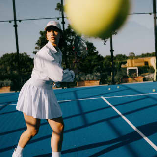 Action shot as a woman in whites hits a ball towards the camera, obscuring the image, in a detail of the blue tennis court.