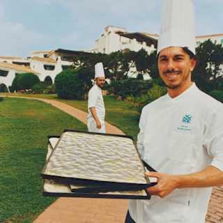 A chef in a white top and toque stands on a garden path with three stacked trays of focaccia dough, as another chef watches.