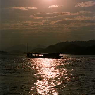 The silhouette of a traditional wooden long boat sails through a trail of light on the Mekong River during a sunset cruise.