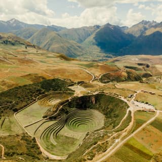 Aerial view of Moray, comprising grassy terraced circular depressions in a patchwork plateau with dramatic mountain backdrop.