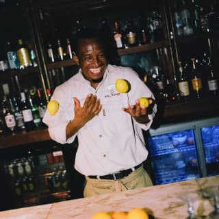 A barman at Eagle Island Lodge laughs as he juggles three lemons behind the marble-top bar, with bottles shelves behind.