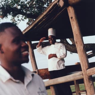 A chef in a toque smiles and salutes from the Fish-Eagle Bar, while a waiter with a tray passes by in soft-focus foreground.