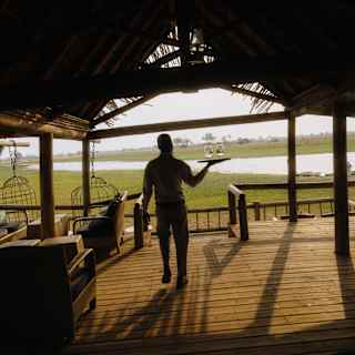 A silhouetted member of staff carries a tray with two glasses across the decked floor of the bar with a view of sunny plains.