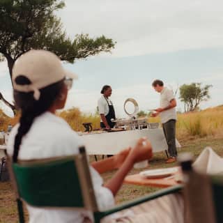 A man helps himself at the al-fresco brunch station, helped by staff, seen over the shoulder of a woman in a white cap.