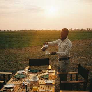 A male staff member pours coffee from a silver jug into a cup at an al-fresco breakfast table as the sun rises.