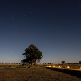 Lantern lit platform over grasslands under a starry sky with sunrise bursting on the horizon