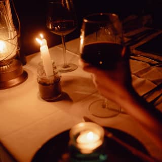 In the darkness of the bar at night, a guest raises a glass of red wine at a table illuminated by two candles and a lamp.