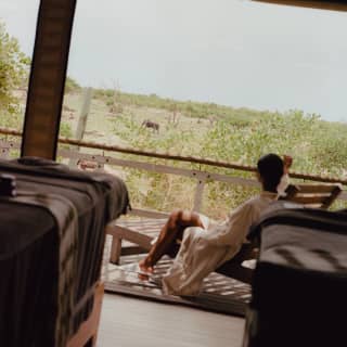 A man in a white robe and slippers reclines in a lounger on the deck of his accommodation, gazing at a passing elephant.