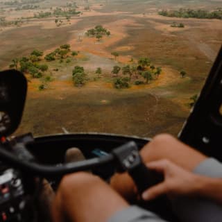 View of the Savannah in patches of parched brown and russet red, studded with trees, seen from a helicopter cockpit.