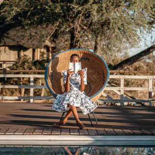 A female guest sits in a round wicker chair reading a book alongside the pool