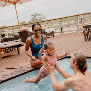 A woman in a blue swimming costume laughs as her daughter jumps into the arms of a man in their private plunge pool.
