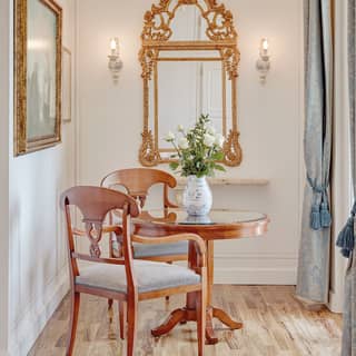 A gilt Rococco mirror hangs in refined dining nook furnished with table and chairs for two. Full height curtains frame a window