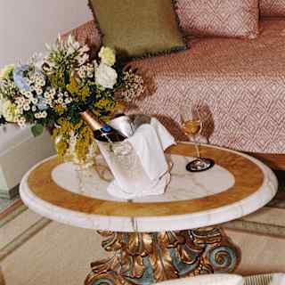 A flower vase and wine bucket holding champagne, with a half-full glass, rest on a table with a marble top and gilt pedestal.