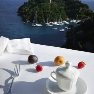 A flotilla of white hulled sailing boats are moored in the bay with the steep, treelined sides of Punta de Coppo rising behind