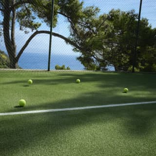 Yellow tennis balls lie behind the white painted baseline of an artificial grass court. Beyond the fencing are stunning sea views