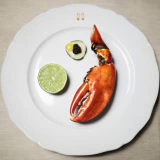 A sunset orange lobster claw is arranged on a white plate with half a lime and a garnish of caviar atop a slice of cucumber