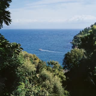 A boat crosses the deep blue sea which meets a lightly clouded horizon in a high-up view between foliage-covered hills.