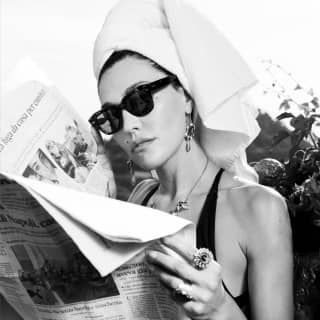 A woman in a black swimsuit and sunglasses reads a newspaper in the sun, her hair wrapped in a white towel turban