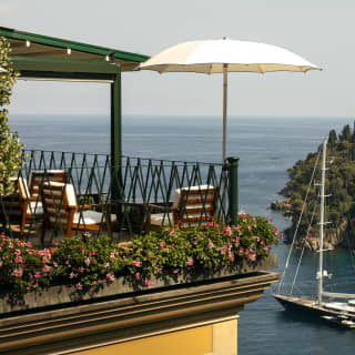 Geraniums fill the edge of the restaurant balcony beside teak chairs with deep cushions. Below a large sail boat is moored