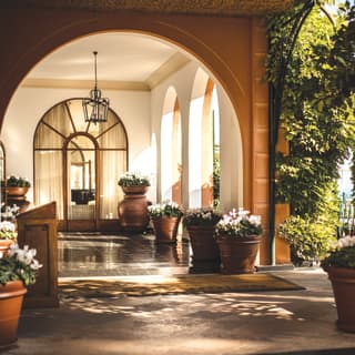 Hotel entrance under a stone arch beyond a garden patio dotted with potted plants