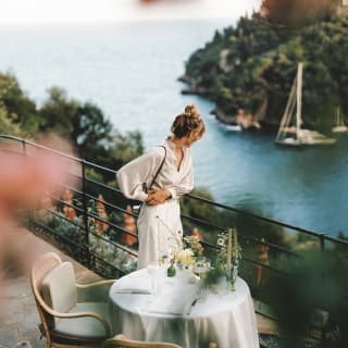A woman in white leans on terrace railings behind a dining table for two, gazing at the dazzling bay, viewed through foliage.
