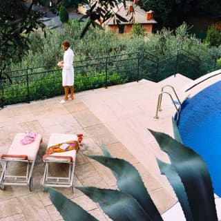 A man in a white robe and slippers stands, looking at the gardens in front of two poolside sun-loungers, seen from above.