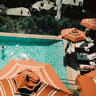 View over orange and white parasols to the pool, where two guests enjoy a sun-kissed swim and others sunbathe at the side.