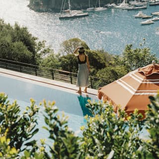 A female guest stands at the edge of the hilltop pool, looking over the bay to where boats huddle along the Punto del Coppo.
