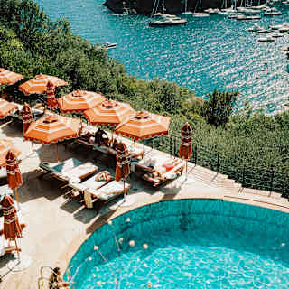 A high-angle image captures guests relaxing by the pool and on loungers shaded by orange parasols, above the glittering bay.