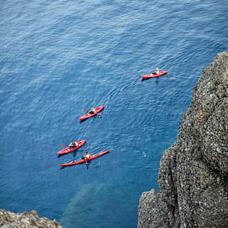 Paddlers in four kayaks explore the steep cliff coastline; their bright red kayaks small against the vastness of the sea