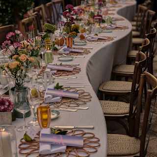 Close-up of a wavy banquet table adorned with pretty flower vases and place settings with wicker mats, napkins and scrolls.