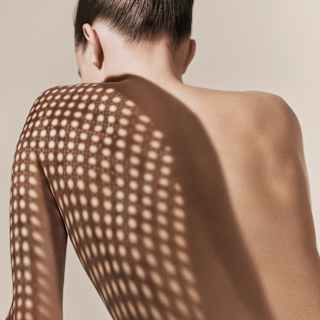 Sun through wicker casts a spotty shadow over the shouder, like a shawl, on the naked back of a woman, seen from behind.