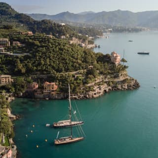 Two single-mast sailing ships float in the spectacular Baia Canone, seen from above, with a backdrop of forested hills.