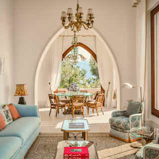 View over a living room with a large blue sofa and two armchairs through a wall arch to a dining area with sublime sea views.