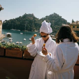 Two women in bathrobes eating and laughing on a balcony, Portofino harbour in the background