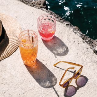 A Cosmopolitan and a Negroni sit on a stone wall overlooking the sea. A straw hat and Jackie O sunglasses lie discarded