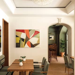 Elegant mid-century sage green chairs sit at polished pale wood tables. An abstract geometric painting hangs on the far wall