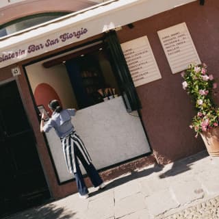 Angled image of a dark-haired waitress standing at the hatch of Gelateria San Giorgio in a striped apron, seen from behind.