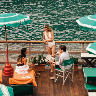 Young people in bathing suits relax on a large balcony with green parasols as the sparkling emerald sea extends far beyond