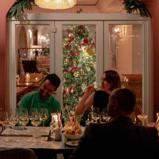 A Christmas tree glows behind glass doors, in front of which a woman sips wine with two male friends at a festive table.